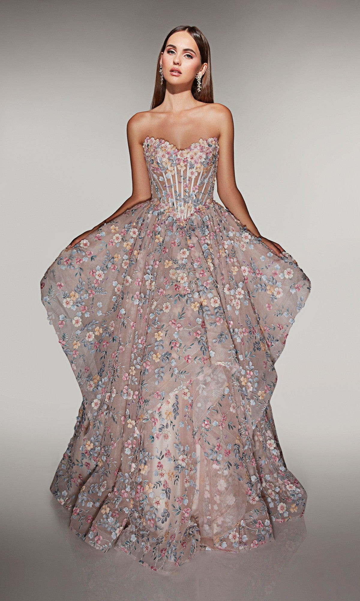 A woman wears the Alyce 62048 long prom dress, featuring a strapless sheer corset bodice and voluminous skirt adorned with floral appliques as she gracefully extends its sides.